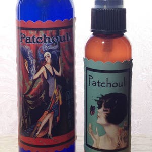 Puede incluir: Dos botellas de vidrio de aceite esencial de pachulí. La botella más grande es azul con una etiqueta de estilo vintage que presenta a una mujer con un vestido rojo. La botella más pequeña es ámbar con una etiqueta verde que presenta a una mujer con una mariposa.