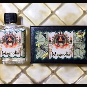 Könnte beinhalten: Eine kleine, durchsichtige Glasflasche mit schwarzem Verschluss und einem Etikett mit der Aufschrift "Magnolia". Die Flasche steht neben einer schwarzen Schachtel mit einem grünen Etikett, auf dem ebenfalls "Magnolia" steht. Das Etikett zeigt eine Frau in einem Kleid im Vintage-Stil und ein Blumenmuster.