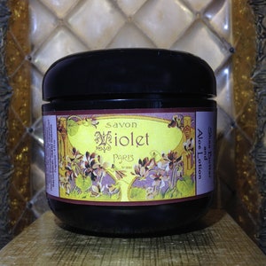 May include: A black jar with a yellow and brown label that reads "Savon Violet Paris" and "Shea Butter and Aloe Lotion". The label features a floral design with purple flowers and green leaves.