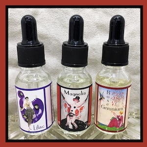 Puede incluir: Tres pequeños frascos de vidrio con goteros y etiquetas. Las etiquetas presentan ilustraciones y texto de estilo vintage. Los frascos están etiquetados "Lilac", "Magnolia" y "Rose Geranium".