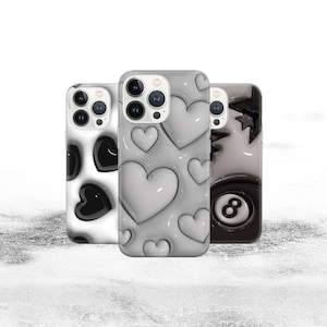 Puede incluir: Tres fundas de teléfono diferentes con varios diseños. La primera funda tiene un patrón de corazón blanco y negro. La segunda funda tiene un patrón de corazón blanco. La tercera funda tiene un diseño de bola 8 blanco y negro.