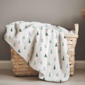 May include: A white blanket with a repeating pattern of green and blue-green watercolor Christmas trees is draped over a woven wicker basket. The blanket is soft and fluffy, with a cozy appearance. The basket has a natural wood color.