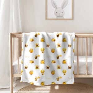 May include: White baby blanket with a repeating pattern of yellow bees and honey jars. The name "Olivia" is printed in gold lettering on the blanket.
