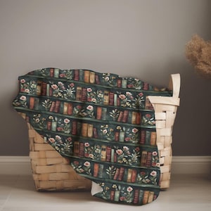 May include: A dark green blanket with a repeating pattern of bookshelves, books, and flowers. The blanket is draped over a woven basket. The books are various colors, and the flowers are pink and white.