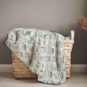 May include: A soft, cream-colored blanket with a watercolor print of bookshelves and potted plants. The design features rows of books in shades of blue and gray, with green foliage. The blanket is draped over a woven basket.