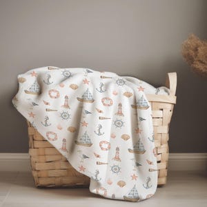 May include: A white nautical-themed blanket with a repeating pattern of sailboats, anchors, lighthouses, and seashells in shades of blue, orange, and brown. The blanket is draped over a woven basket.