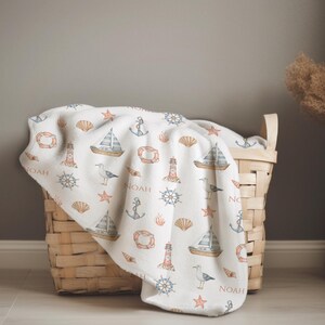 May include: A white baby blanket with a nautical theme. The blanket features a repeating pattern of sailboats, anchors, lighthouses, seagulls, seashells, and starfish. The name "Noah" is printed in red on the blanket.