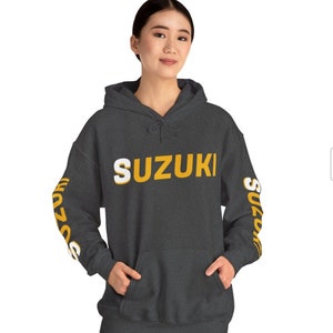 May include: A dark grey hooded sweatshirt with the word "SUZUKI" printed in white and yellow on the front. The word "SUZUKI" is also printed on the sleeves in yellow and white.