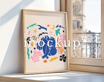 May include: A framed print with a colorful abstract design featuring a blue figure, stars, and flowers. The frame is made of natural wood and has a light brown finish. The print is displayed on a white surface next to a window.