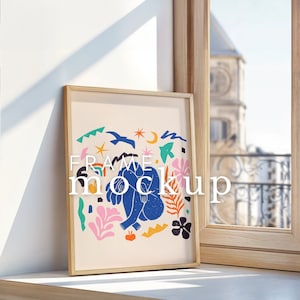 Puede incluir: Una impresión enmarcada con un diseño abstracto colorido que presenta una figura azul, estrellas y flores. El marco está hecho de madera natural y tiene un acabado marrón claro. La impresión se muestra en una superficie blanca junto a una ventana.