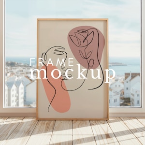 May include: A framed print with a minimalist line drawing of a face and a flower. The print is on a beige background with pink and peach accents. The frame is light wood.