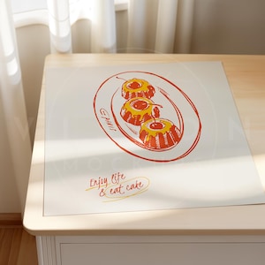 K&ouml;nnte beinhalten: Ein wei&szlig;er Platzdeckchen mit einer handgezeichneten Illustration von drei gelben und roten Gugelhupfkuchen mit Kirschen darauf. Das Platzdeckchen tr&auml;gt den Text "Enjoy life & eat cake" in Orange.