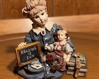 Könnte beinhalten: Eine Figur aus zwei Mädchen, eines hält eine Tafel, auf der "Reading is Fun" steht, und das andere sitzt auf einem Stapel Bücher. Die Mädchen sind in blaue und rote Kleider gekleidet und haben blondes Haar.