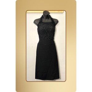 以下が含まれることがあります： マネキンに展示された黒いホルターネックドレス。トップスは質感のあるフリルデザインで、スカートはシンプルな膝丈スタイルです。ドレスは無地の黒色です。