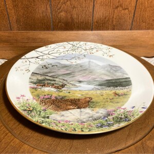 May include: A white porcelain plate with a gold rim features a scenic landscape painting of a mountain range, a river, and two grouse birds. The plate is on a wooden surface.