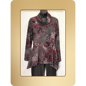 May include: A long-sleeved tunic top with a cowl neck, featuring a floral pattern in shades of gray, burgundy, and black. The tunic has an asymmetrical hemline and is displayed on a mannequin. The top is paired with dark gray pants.