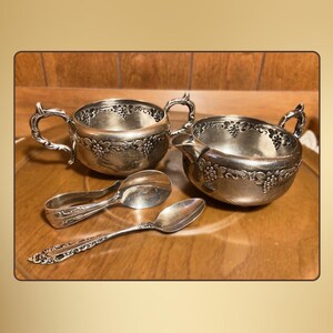 Könnte beinhalten: Silbernes Teeservice mit verzierten Griffen und Weinreben-Details. Enthält eine Zuckerdose, einen Sahnekännchen und zwei Löffel. Das Set wird auf einer Holzoberfläche präsentiert und zeigt sein elegantes Design und seinen Vintage-Charme.