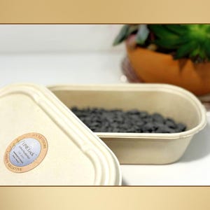May include: An open, beige rectangular container with dark gray contents, next to a matching container with a lid. The lid has a circular sticker with text. A potted plant is visible in the background. The containers appear to be made of a natural material.