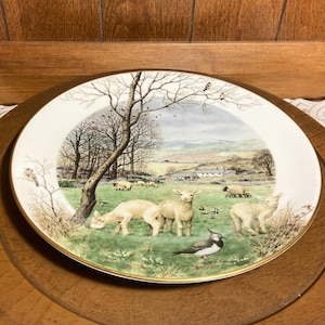 May include: A white ceramic plate with a painted scene of a rural landscape with sheep and birds. The plate has a gold rim and is on a wooden surface.