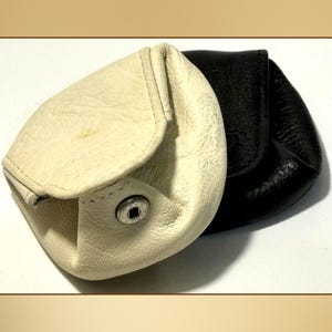 Pode incluir: Duas bolsas de moedas de couro, uma creme e outra preta. A bolsa creme está aberta, mostrando o interior, e tem um fecho de pressão. Ambas as bolsas têm formato arredondado e superfície texturizada.