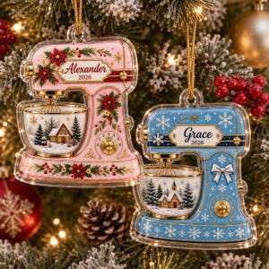 Könnte beinhalten: Zwei flache, transparente Acryl-Ornamente in Form von Küchenmaschinen. Das rosa Modell zeigt den Namen Alexander und das Jahr 2026 mit Weihnachtssternen. Das blaue Modell zeigt den Namen Grace und das Jahr 2026 mit Schneeflocken und Schleife.