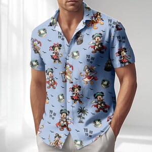 May include: Light blue button-down shirt with a pirate-themed pattern featuring cartoon characters. The design includes images of Mickey and Minnie Mouse, Donald Duck, and Goofy dressed as pirates, along with pirate ships and the text "YO HO".
