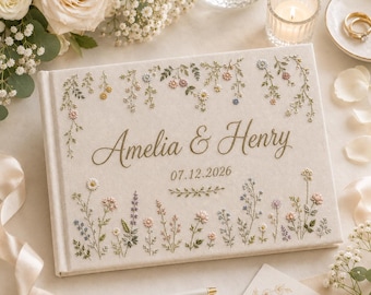 May include: A cream-colored fabric photo album featuring embroidered wildflowers in shades of pink, blue, yellow, and green. The cover displays the names Amelia & Henry in gold script, followed by the date 07.12.2026 and a small floral motif.