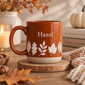 May include: A burnt orange ceramic mug featuring the name Hazel printed in white text above a row of white botanical leaf illustrations. The mug has a smooth, glossy finish on the upper portion and a matte, light beige base with a curved handle.