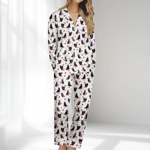 Könnte beinhalten: Weißes Pyjama-Set mit einem Muster aus schwarzen Katzen und roten Herzen. Das langärmlige Oberteil hat eine Knopfleiste und einen Reverskragen. Die Hose hat eine lockere Passform. Der Pyjama ist aus einem weichen, leichten Stoff gefertigt.