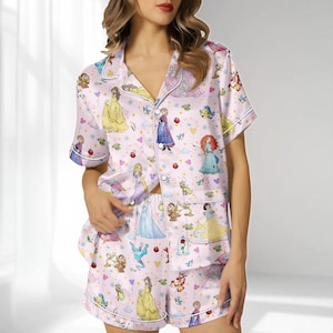 Könnte beinhalten: Rosafarbenes Pyjama-Set mit einem Muster von Cartoon-Prinzessinnen. Das kurzärmelige Oberteil und die Shorts haben weiße Paspeln. Das Set enthält ein Hemd mit Knöpfen, einem Reverskragen und einer Tasche.