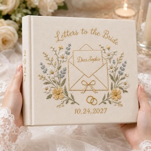 May include: A cream-colored fabric-bound book titled Letters to the Bride with gold embroidered text, a central envelope graphic labeled Dear Sophia, floral embroidery in blue, white, and yellow, two gold rings, and the date 10.24.2027.