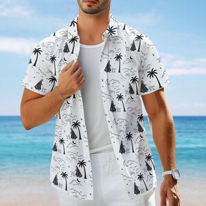Könnte beinhalten: Weißes kurzärmeliges Hemd mit einem schwarzen und grauen tropischen Muster mit Palmen, Figuren, Wolken und Monden. Das Hemd wird offen über einem weißen Unterhemd getragen und mit einer weißen Hose kombiniert, was auf ein Urlaubsthema hindeutet.