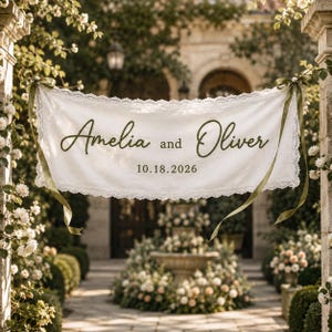 May include: A white fabric banner with lace trim featuring the dark green text Amelia and Oliver and the date 10.18.2026. The banner is suspended by olive green ribbons tied at each end, displaying a script font for the names.