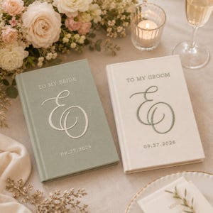 Könnte beinhalten: Zwei Hardcover-Bücher mit Stoffeinband. Das salbeigrüne Buch zeigt den weißen gestickten Text TO MY BRIDE, einen großen geschwungenen Buchstaben E und das Datum 09.27.2026. Das cremefarbene Buch zeigt den salbeigrünen Text TO MY GROOM, ein E und 09.27.2026.