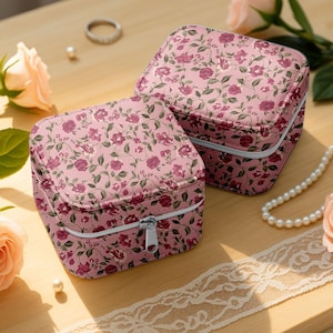 Könnte beinhalten: Zwei rosa Schmuckkästchen mit Blumendruck und silbernen Reißverschlüssen. Die quadratischen Boxen weisen ein Muster aus burgunderfarbenen Blumen und grünen Blättern auf. Die Boxen stehen auf einer hellbraunen Holzoberfläche, mit einer Perlenkette und einem Ring im Hintergrund.