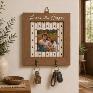 Könnte beinhalten: Ein quadratischer Schlüsselhalter aus Holz zur Wandmontage mit einem Fotorahmen, der einen weißen Rand mit kleinen roten Äpfeln aufweist. Der Rahmen zeigt ein Foto von zwei Personen und ist mit dem Text Lucas und Harper graviert. Zwei Metallhaken halten Schlüssel.