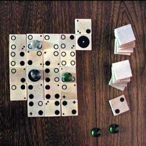 May include: A wooden game board with black and white circles and a plus sign. There are also green and white marbles on the board. The board is surrounded by wooden tiles with black and white circles.