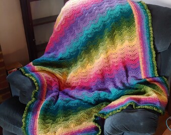 May include: A colorful, handmade crochet blanket draped over a dark blue armchair. The blanket features a vibrant rainbow of colors, including pink, yellow, green, blue, and purple. The edges are finished with a scalloped green border, adding a decorative touch to the cozy blanket.