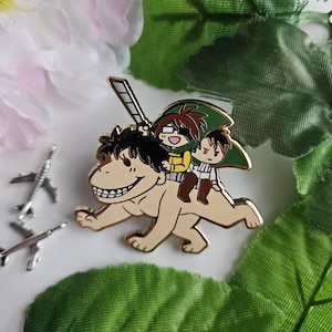 Könnte beinhalten: Ein goldfarbener Emaille-Pin mit Figuren aus der Anime-Serie Attack on Titan. Der Pin zeigt drei Charaktere, die auf einem Titanen reiten, von denen einer ein Schwert hält. Der Pin ist von grünen Blättern und rosa Blüten umgeben.
