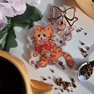 Op de afbeelding: Een heldere acryl kattenbedel met oranje en bruine strepen, een rode strik en koffiebonen. Een roségouden sleutelhanger in de vorm van een strik en een kopje koffie zijn ook zichtbaar. Een gele mok staat op de voorgrond.