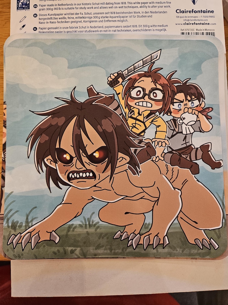Puede incluir: Una alfombrilla de rat&oacute;n rectangular con una colorida ilustraci&oacute;n de estilo anime. La obra de arte representa a tres personajes montados sobre la espalda de una gran figura monstruosa. El fondo es un campo de hierba. La alfombrilla tiene un logotipo de Clairefontaine.