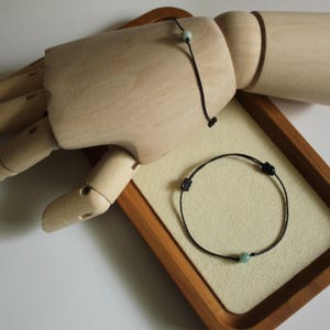 Könnte beinhalten: Zwei schwarze Kordelarmbänder mit kleinen, runden, hellgrünen Perlen. Eines liegt auf einer Holzhand, das andere auf einer beigen Oberfläche in einem Holztablett. Die Armbänder sind schlicht und minimalistisch.