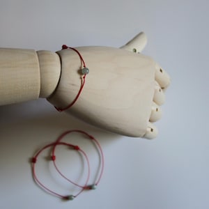 Könnte beinhalten: Ein rotes Armband mit einer kleinen, runden, grauen Perle wird auf einer Holzhand gezeigt. Zwei weitere Armbänder mit ähnlichem Design sind darunter zu sehen. Die Armbänder sind schlicht und minimalistisch.