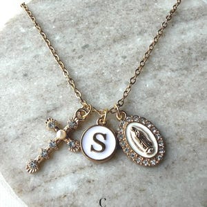 May include: Gold-toned necklace featuring a cross pendant with clear crystals, a round charm with the letter "S" on a white background, and an oval pendant with a religious figure. The necklace is displayed on a marble surface.