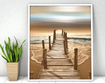 May include: A framed wall art print depicting a wooden pier extending into the ocean at sunset. The scene features a sandy beach, gentle waves, and a soft sky with golden and blue hues. A green potted plant sits to the left of the frame.