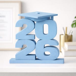 May include: A light blue decorative tabletop sign featuring the numbers 2026 stacked in two rows. The top of the number 2 is topped with a flat, square graduation cap with a small button in the center, all rendered in a solid matte light blue finish.
