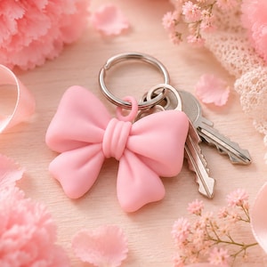 May include: A light pink bow keychain attached to a silver metal split ring with two silver keys. The bow has a smooth, matte finish and is centered among soft pink carnations, delicate baby's breath flowers, and light pink ribbon accents.