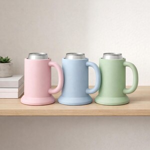 May include: Three pastel-colored can coolers in pink, blue, and green, each with a silver can inside. The coolers have a mug-like design with a handle and a wide base, sitting on a wooden shelf.