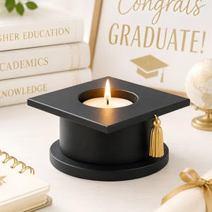 May include: A black graduation cap shaped candle holder with a gold tassel, featuring a lit white tea light candle in the center. The cap is positioned near a stack of books labeled Higher Education, Academics, and Knowledge, a globe, and a diploma.