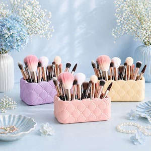 May include: Three makeup brush holders in pastel colors: lavender, pink, and yellow. Each holder is filled with various makeup brushes. The holders have a quilted design. The background features flowers in vases.